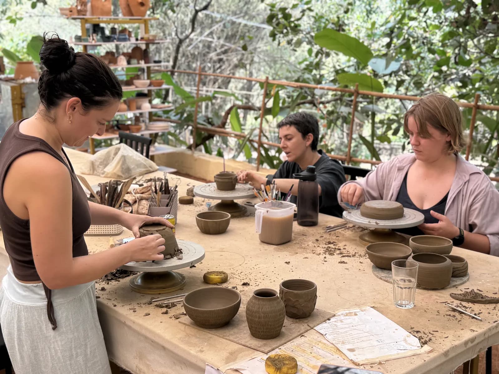 Ceramic students shaping clay and finished vessels at an outdoor Dharamkot Studio class.