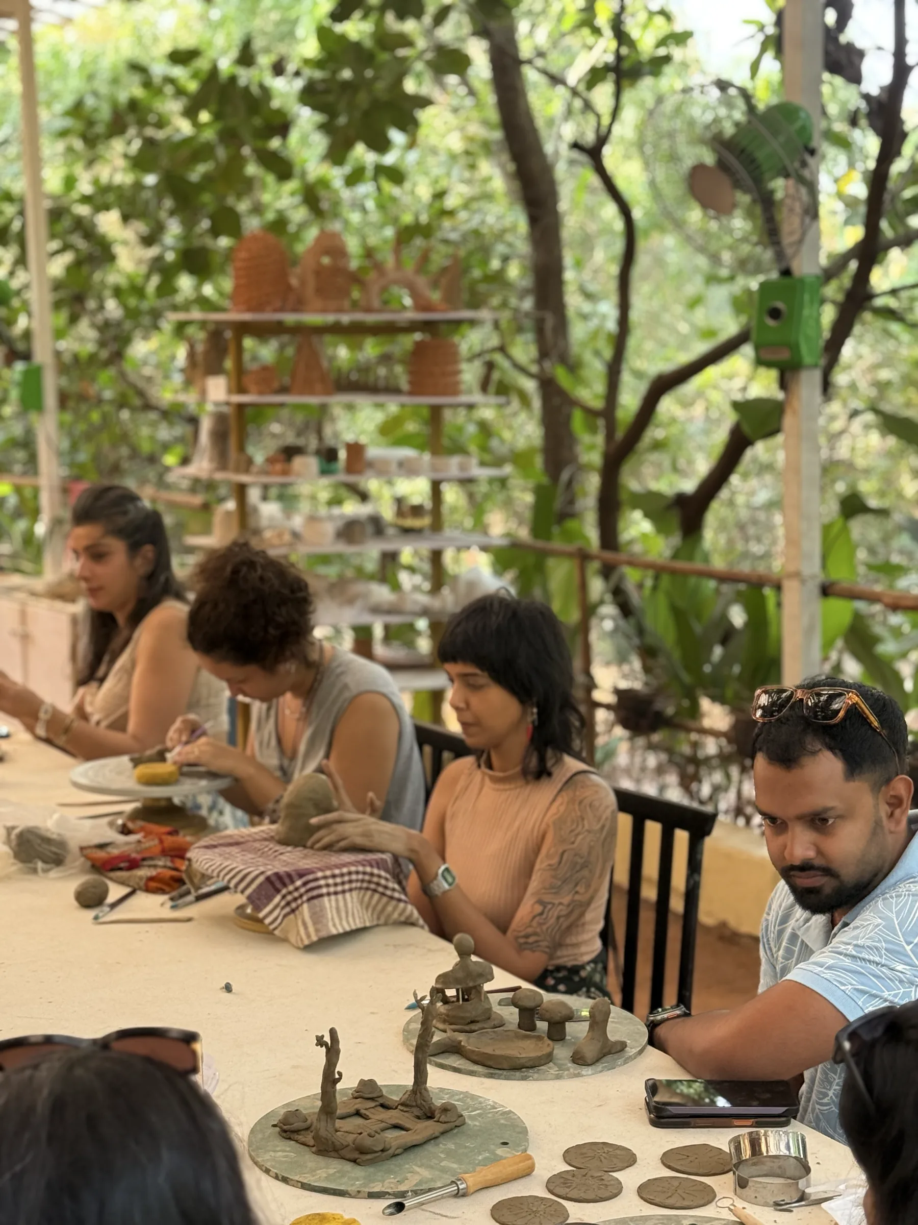Students handbuilding clay at a shaded studio table with greenery around the Goa pottery space.