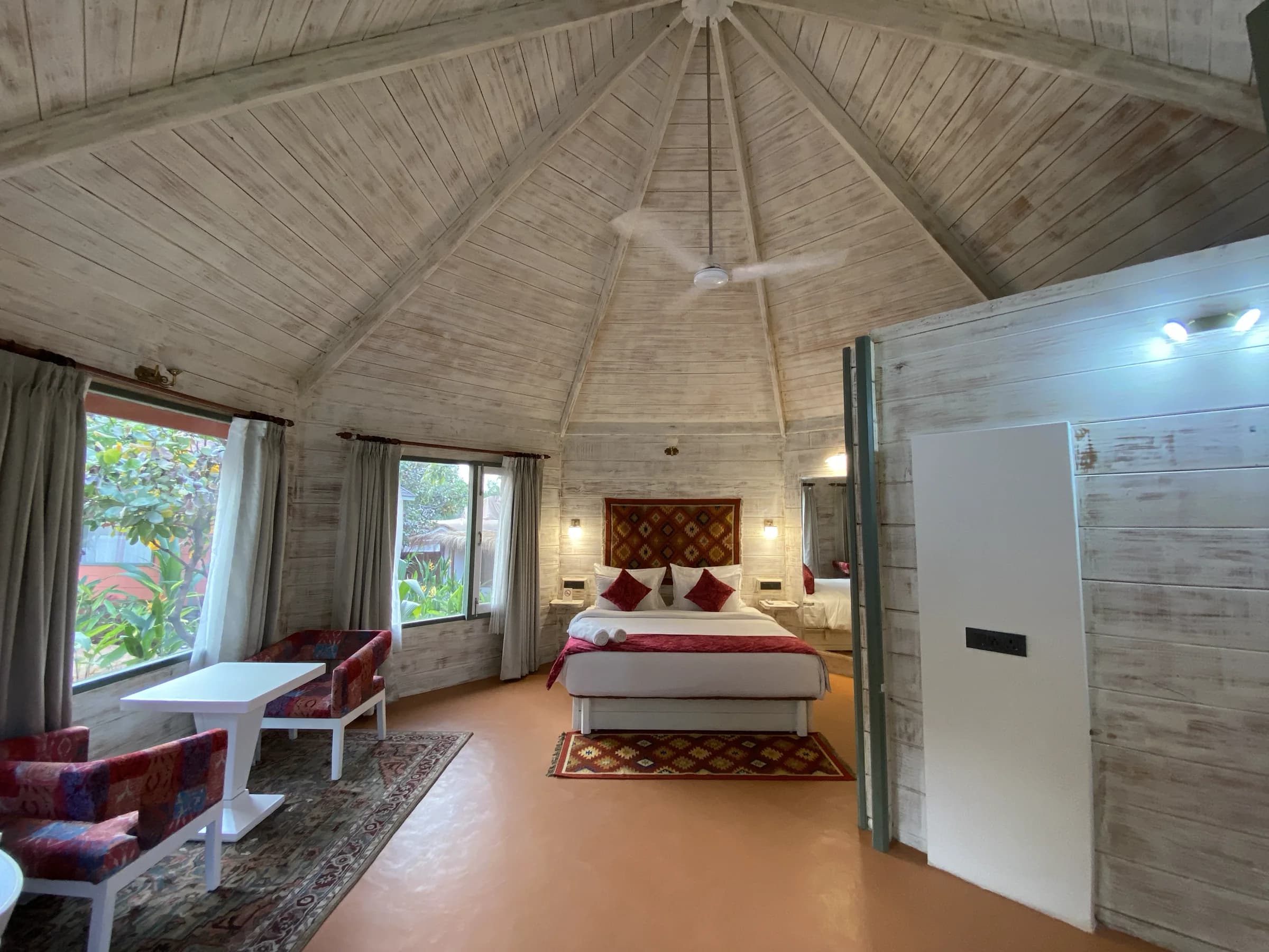A bright wooden cottage bedroom with a neatly made bed, chairs, and large windows at the Goa retreat.