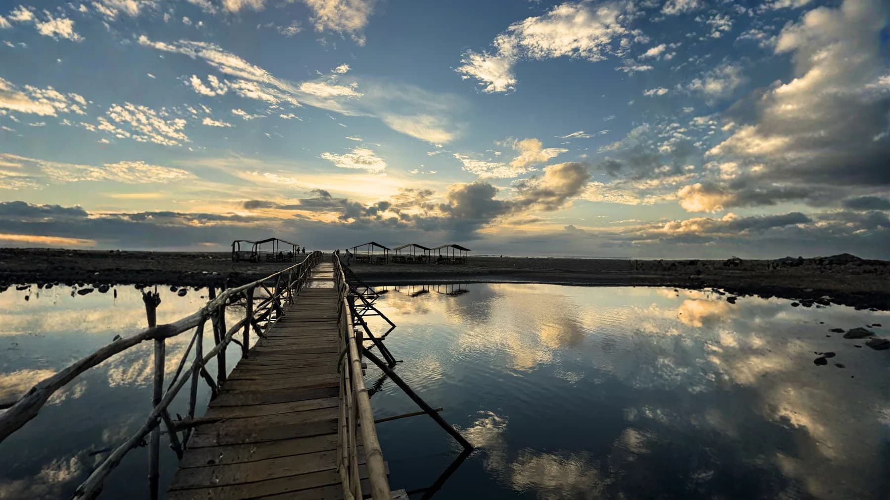 A wooden boardwalk leading toward Mandrem beach under a dramatic blue sky reflected in shallow water.