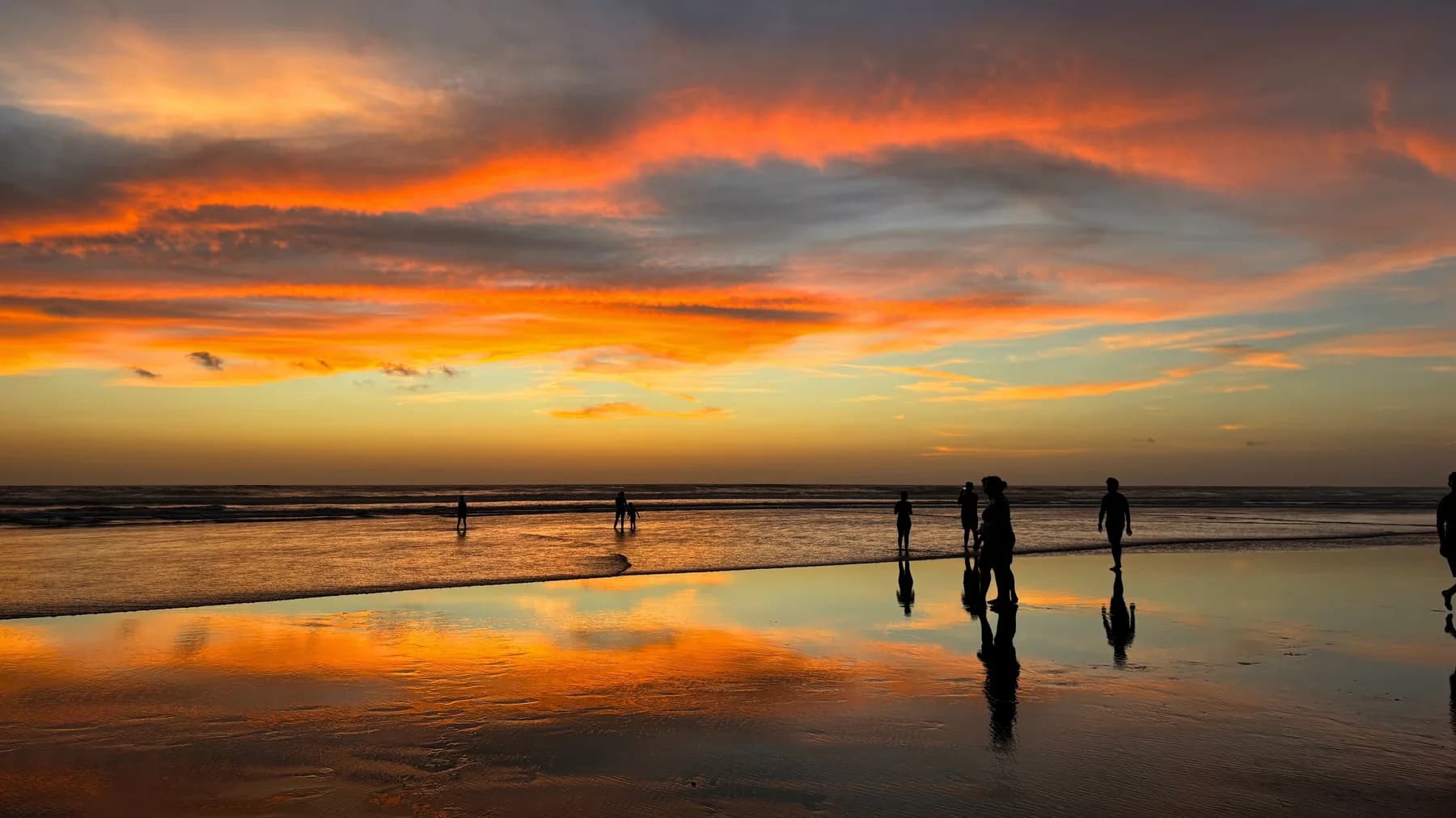 A wide sunset over Mandrem beach with orange clouds, sea reflections, and people walking along the shoreline in Goa.
