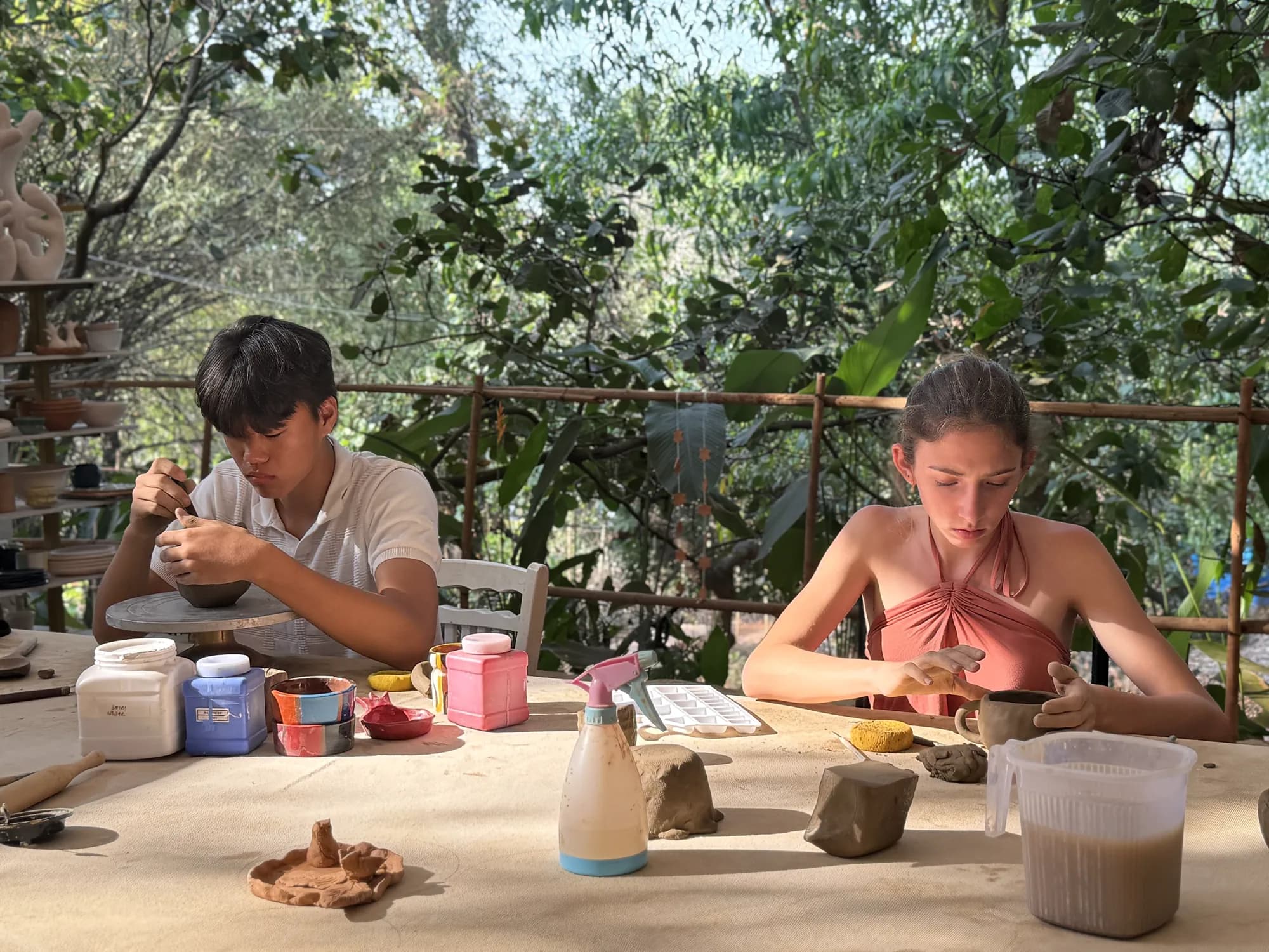 Two young participants quietly shaping bowls at a sunlit clay table in the open-air Goa studio.